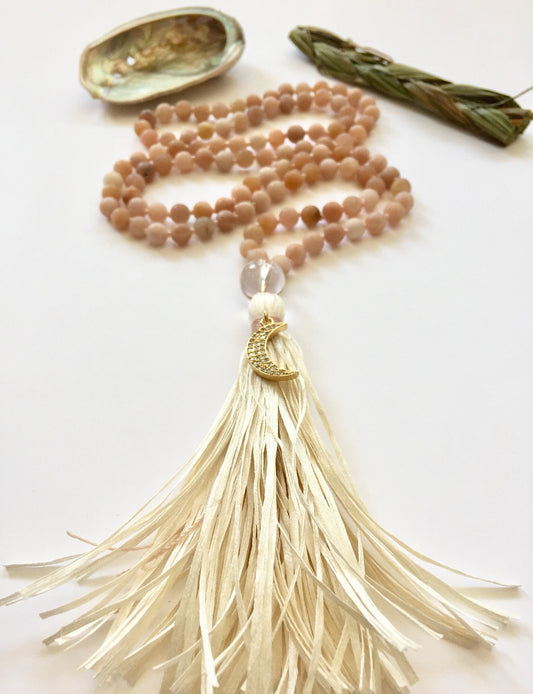 Peach sunstone mala with a cream raffia tassel, an abalone shell and a sage bundle laid above it