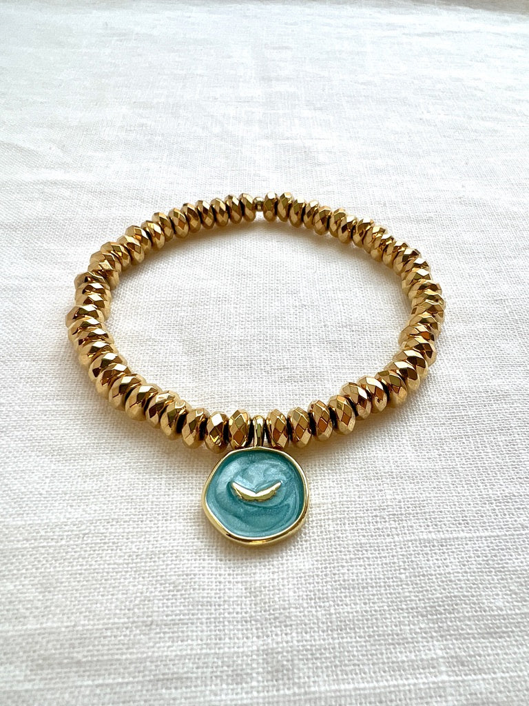 Earth Angel bracelet of faceted gold hematite rondelles with an aqua enamel angel wings charm on cream linen