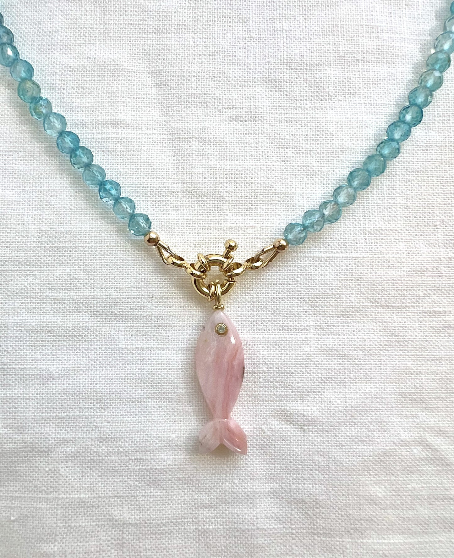 Carved pink opal fish pendant on a faceted blue topaz strand with a gold filled sailor clasp, close view on white linen