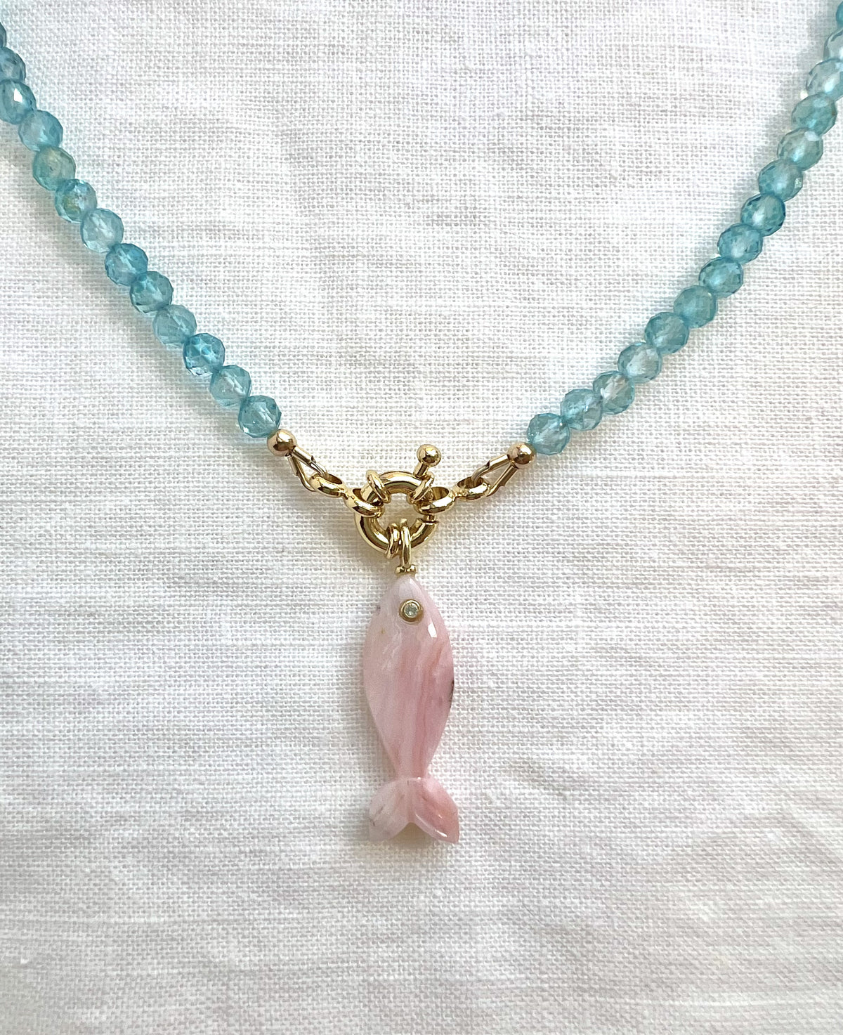 Carved pink opal fish pendant on a faceted blue topaz strand with a gold filled sailor clasp, close view on white linen