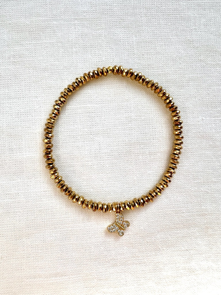 Dainty gold hematite stretch bracelet with a sparkling pave butterfly charm, seen from above on linen