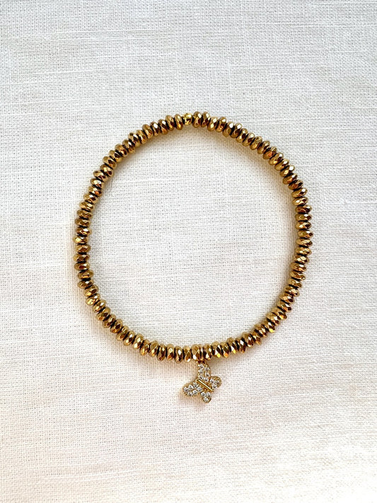 Dainty gold hematite stretch bracelet with a sparkling pave butterfly charm, seen from above on linen