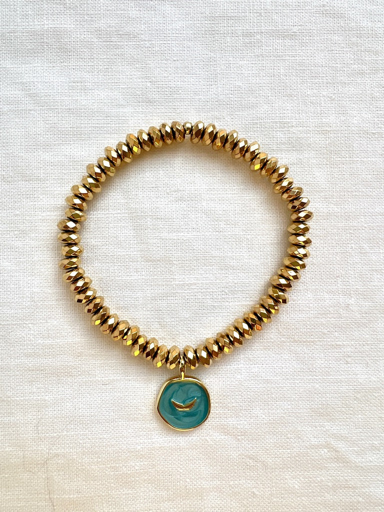 Gold hematite charm bracelet with a teal enamel angel wings charm, seen from above on linen