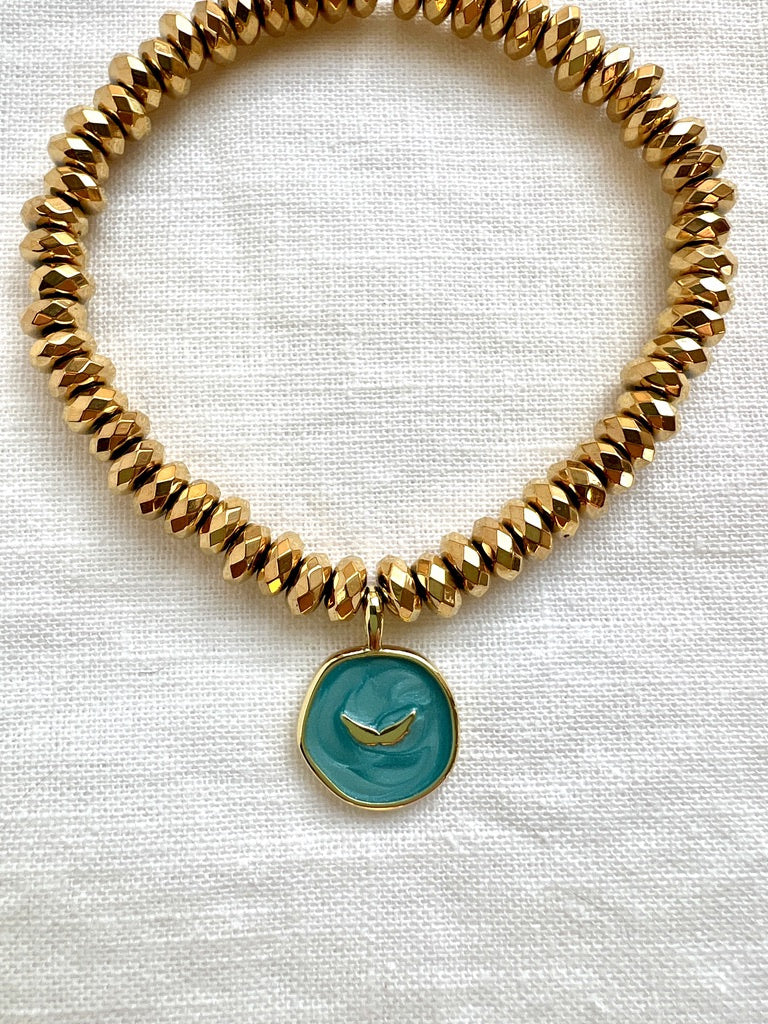 Close up of the aqua enamel charm with raised gold angel wings on a gold hematite bracelet