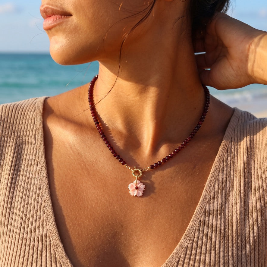 Model wearing the garnet choker with its carved pink shell flower pendant and gold filled sailor clasp at the collarbone, ocean behind her