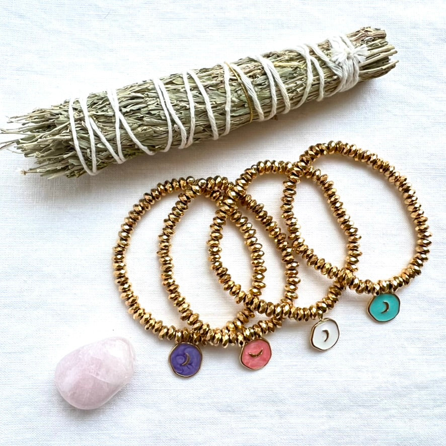 Enamel moon charm bracelets in four colors styled with a sage bundle and a rose quartz stone on white linen