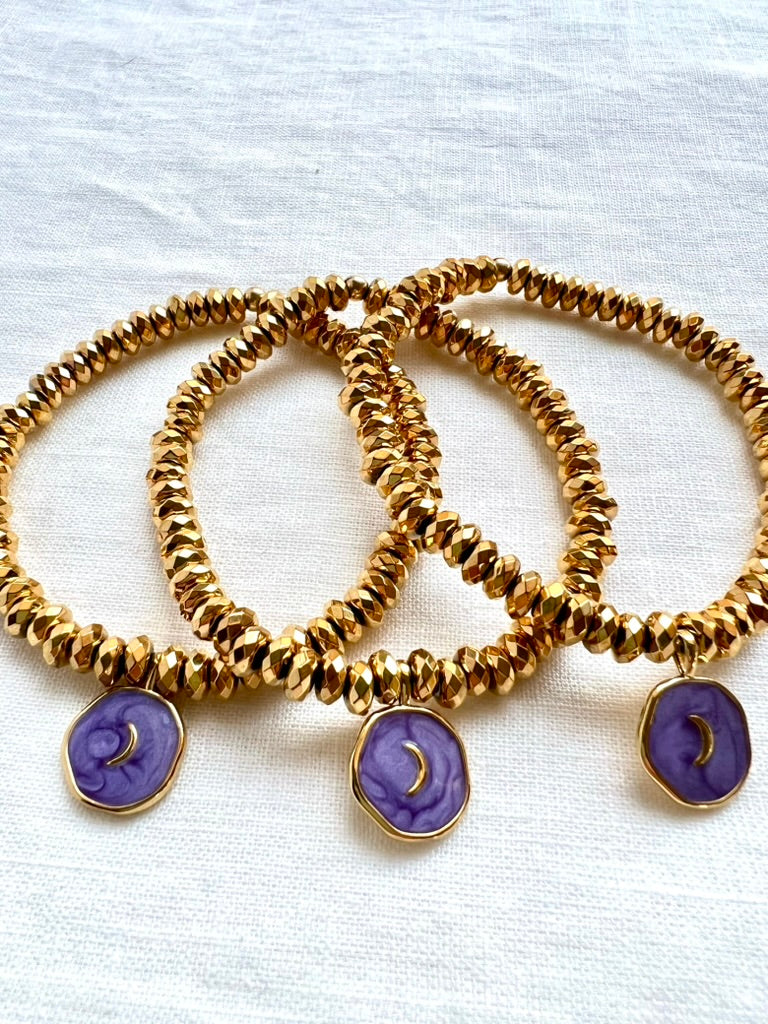 Three purple enamel moon charm bracelets on faceted gold plated hematite beads, stacked on white linen