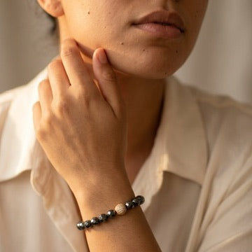 Woman in a cream shirt resting her hand at her chin, wearing the faceted hematite bracelet with a gold pave bead