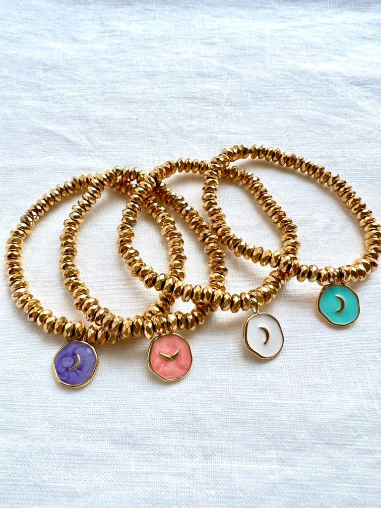 Four enamel moon charm bracelets in purple, pink, white and turquoise on faceted gold plated hematite beads, arranged on white linen