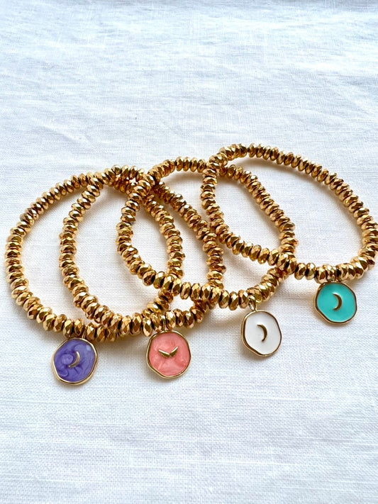 Four enamel moon charm bracelets in purple, pink, white and turquoise on faceted gold plated hematite beads, arranged on white linen