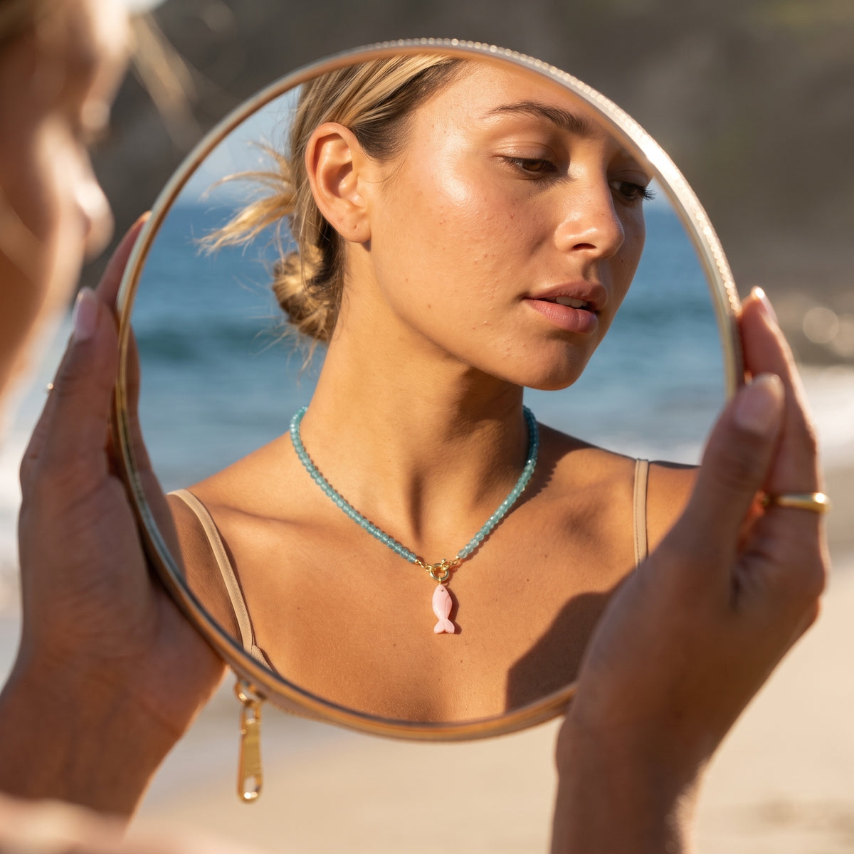 Model wearing the blue topaz choker with its carved pink opal fish pendant, seen in the reflection of a round hand mirror held up at the beach