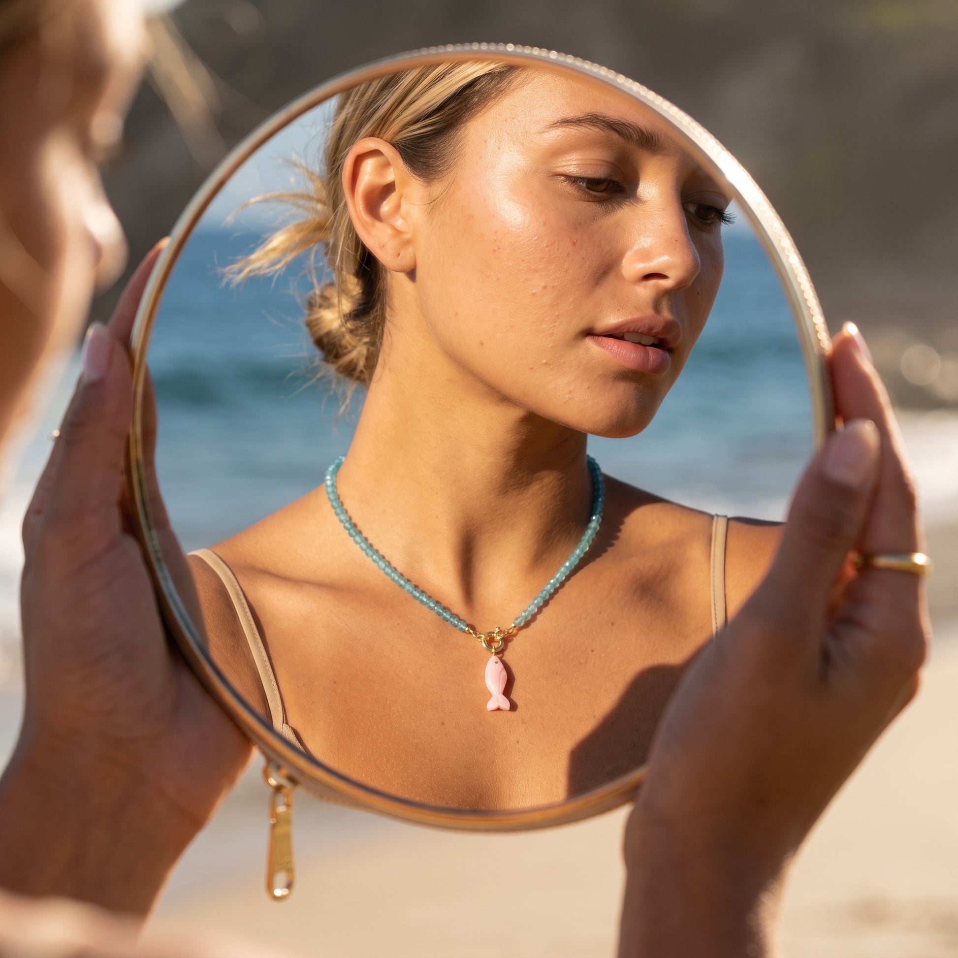 Model wearing the blue topaz choker with its carved pink opal fish pendant, seen in the reflection of a round hand mirror held up at the beach