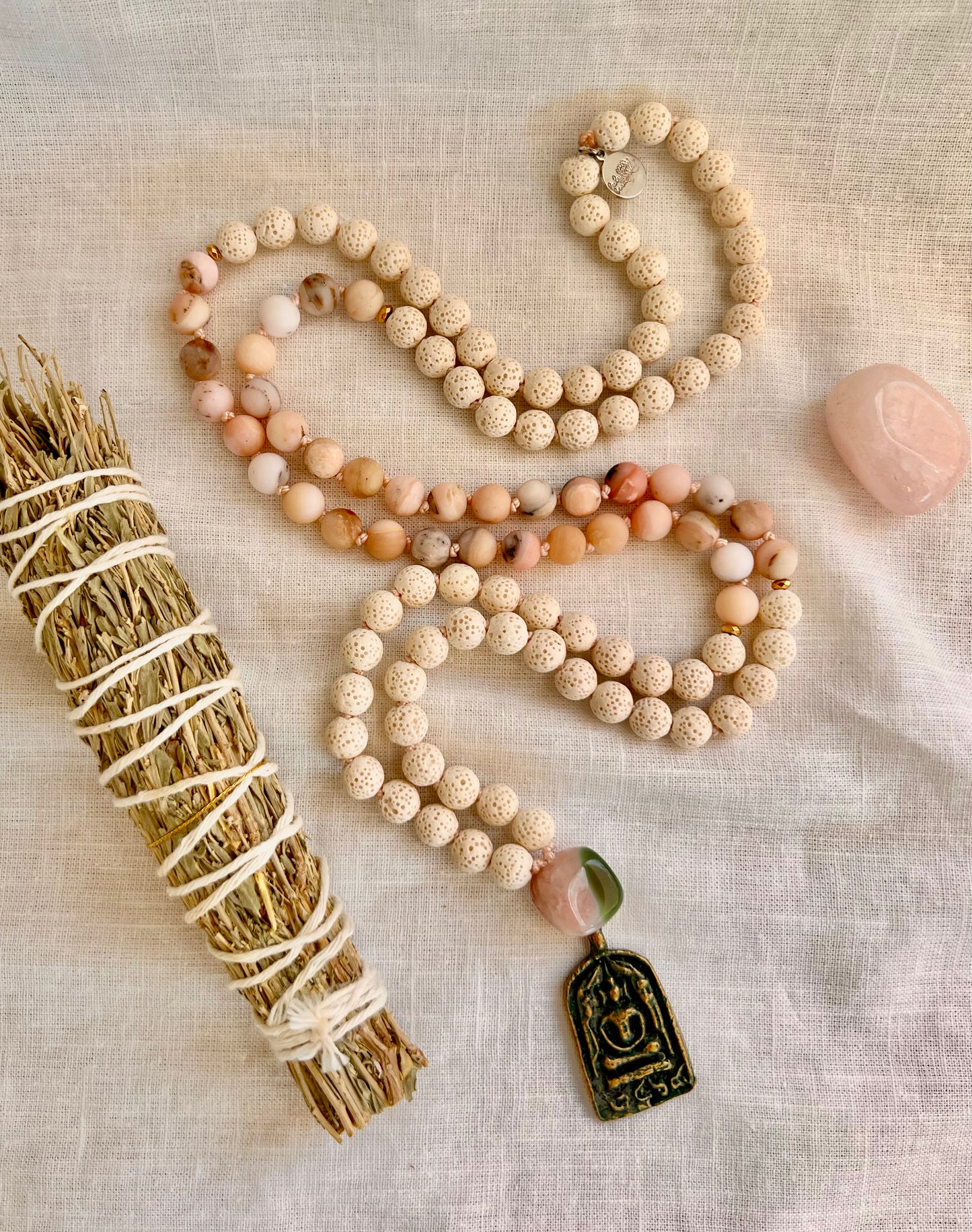Blooming Grace Mala necklace made of white lava and Peruvian pink opal beads with a patina brass Buddha amulet, laid on linen beside a rose quartz stone and white sage bundle