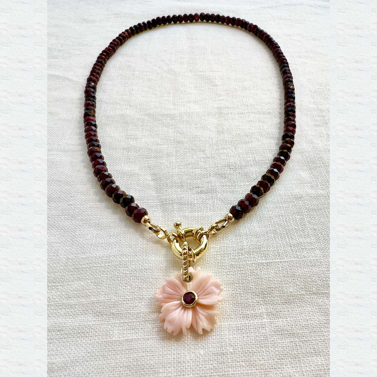 Garnet beaded choker necklace with a carved pink shell flower pendant, angled view on white linen