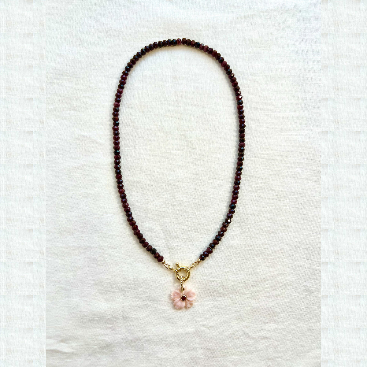 Full view of a faceted garnet rondelle choker necklace with a pink conch shell flower pendant on white linen