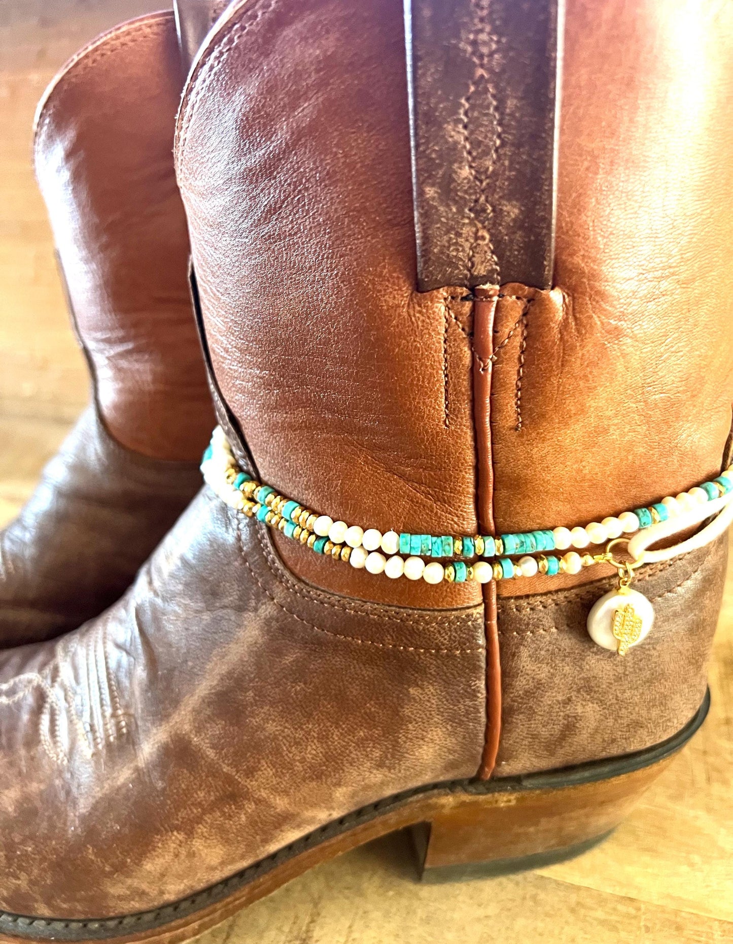 The turquoise and pearl beaded band wrapped around the top of a brown leather cowboy boot, worn as a boot wrap with the gold charm at the front.