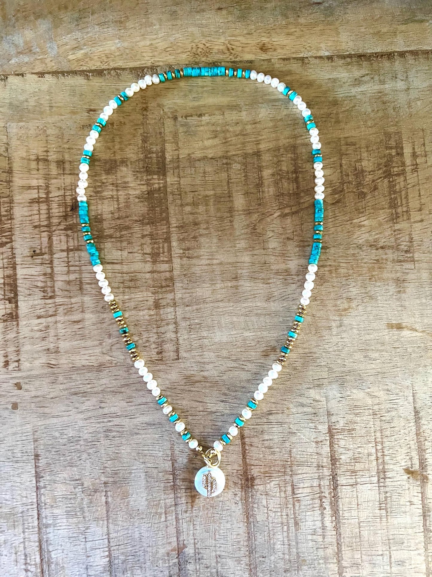 Arizona turquoise, white freshwater pearl and gold hematite necklace laid flat on weathered wood, with the pave cactus charm hanging at the center.