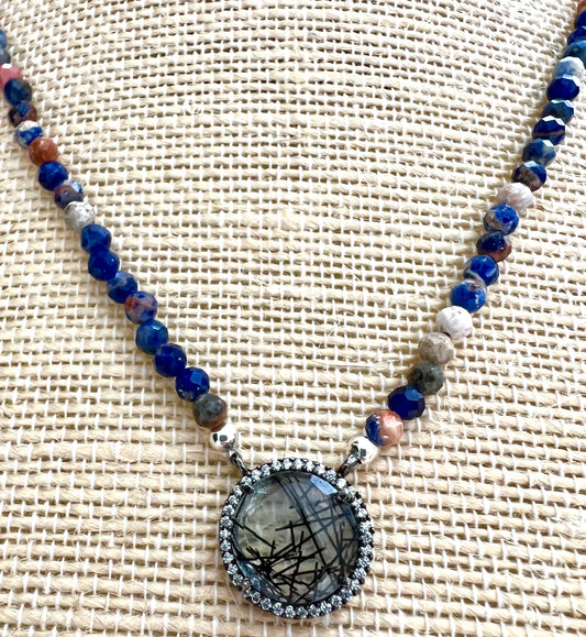 Painted Dusk necklace displayed on woven linen, faceted sodalite beads with a rutilated quartz pendant framed by a halo of cubic zirconia