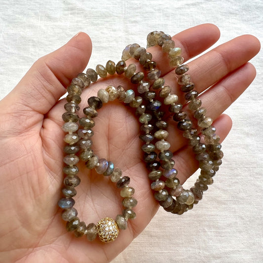 A close-up of a hand holding a double-strand beaded necklace made of faceted gray-green labradorite beads with a small gold pavé accent bead, set against a light beige fabric background.