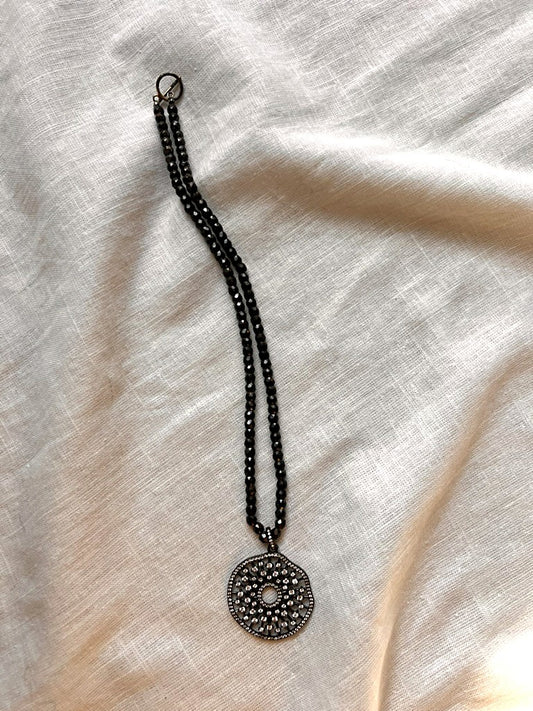 Full view of the Midnight Mandala necklace, a 16 inch strand of faceted black zirconia beads with a dark gunmetal pavé cubic zirconia mandala medallion, laid on cream linen