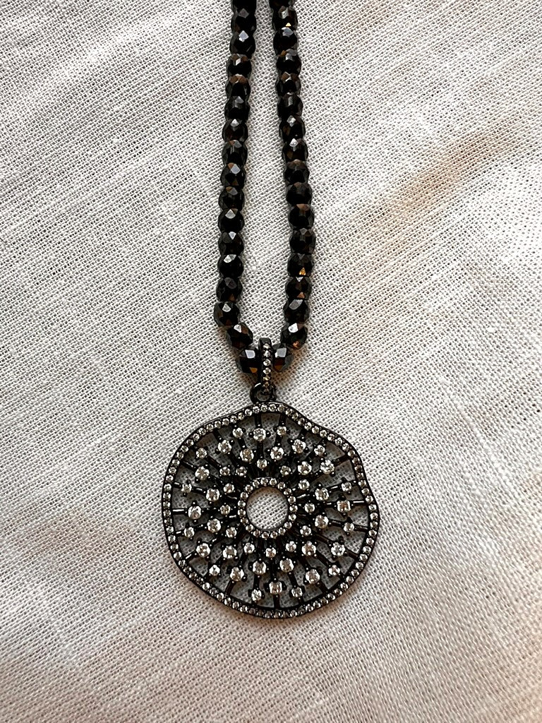 Close up of the 1.5 inch openwork mandala medallion set with sparkling pavé cubic zirconia in a dark gunmetal finish, hanging from faceted black zirconia beads