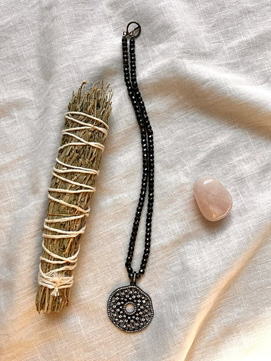 Midnight Mandala necklace with faceted black zirconia beads and a gunmetal pavé cubic zirconia medallion, styled on cream linen with a sage bundle and rose quartz stone