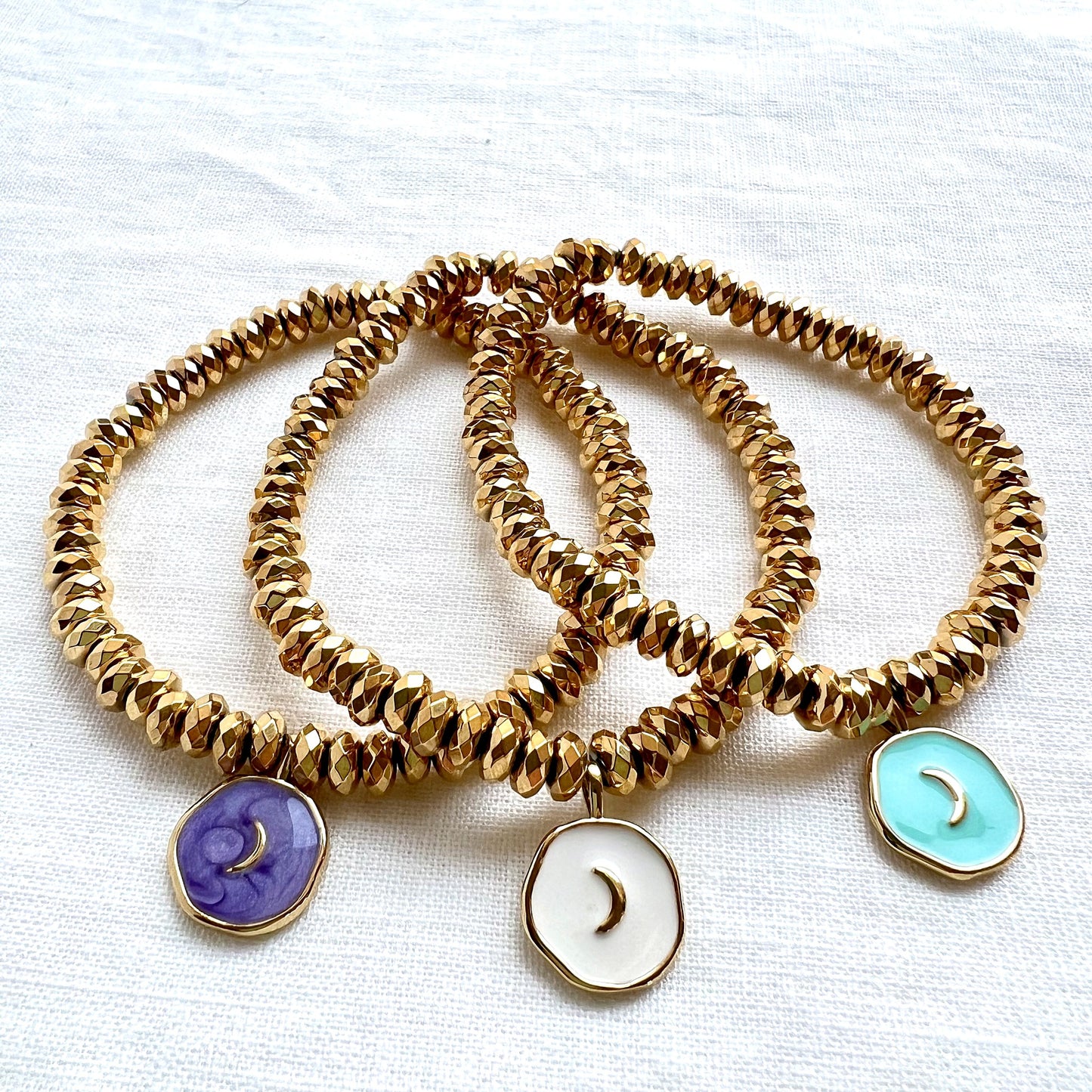 Three enamel moon charm bracelets in purple, white and turquoise on faceted gold plated hematite beads, angled on white linen