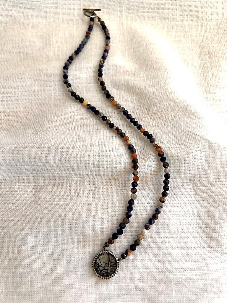 Full view of the Painted Dusk necklace, a 17.5 inch strand of faceted 4mm sodalite beads with a natural rutilated quartz pendant encircled by cubic zirconia, laid on cream linen