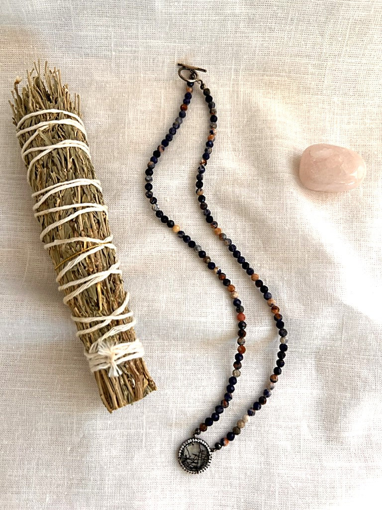 Painted Dusk sodalite necklace with rutilated quartz pendant and sterling silver toggle clasp, arranged on linen beside a sage bundle and rose quartz