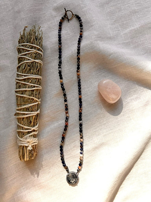 Painted Dusk necklace of faceted 4mm sodalite beads in indigo, terracotta and cream with a rutilated quartz pendant, styled on cream linen with a sage bundle and rose quartz stone