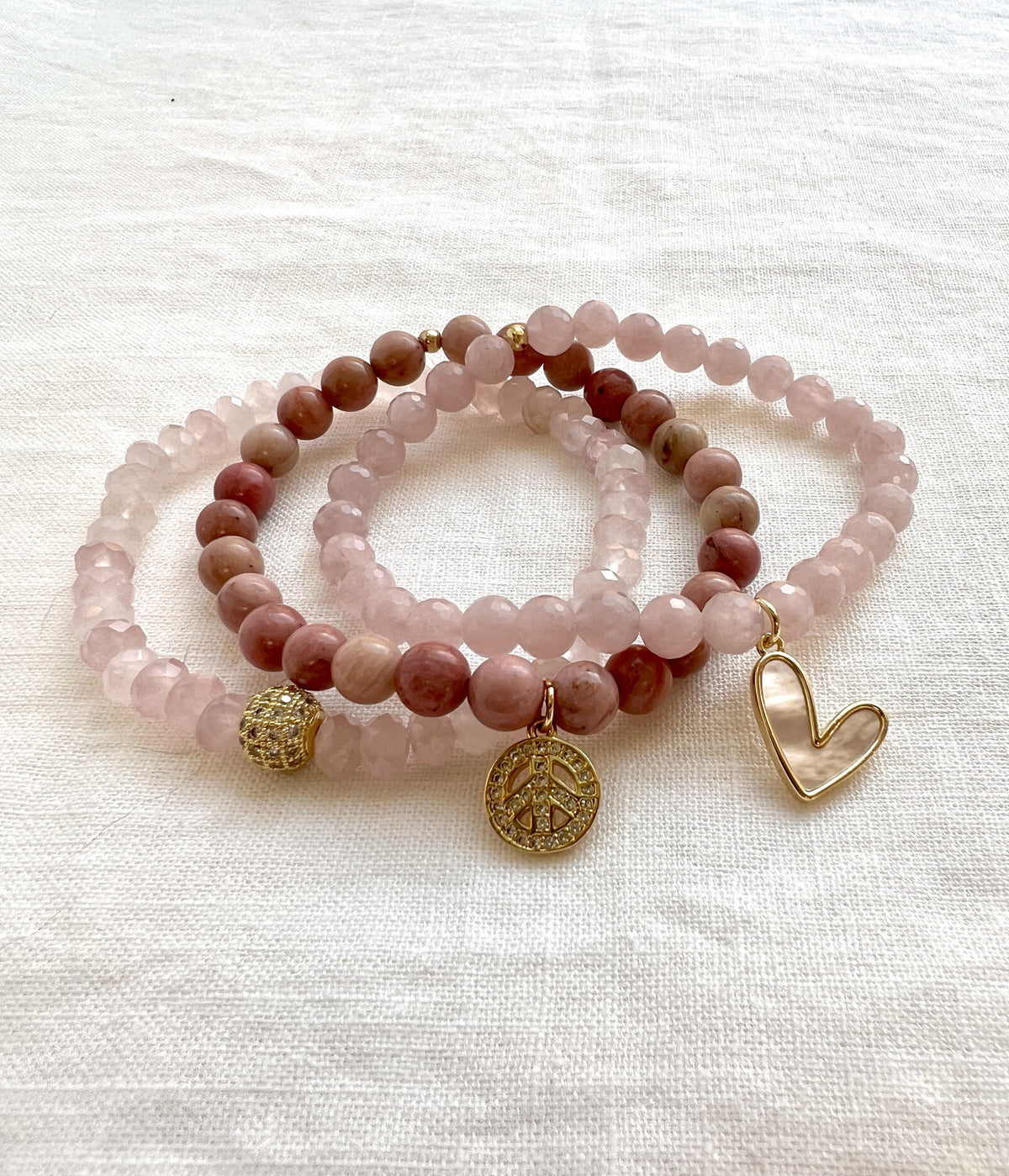 Peace and Love bracelet stack, three rose quartz and rhodonite beaded stretch bracelets on white linen with a gold pave CZ bead, a peace charm and a mother of pearl heart