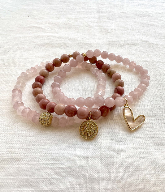 Peace and Love bracelet stack, three rose quartz and rhodonite beaded stretch bracelets on white linen with a gold pave CZ bead, a peace charm and a mother of pearl heart