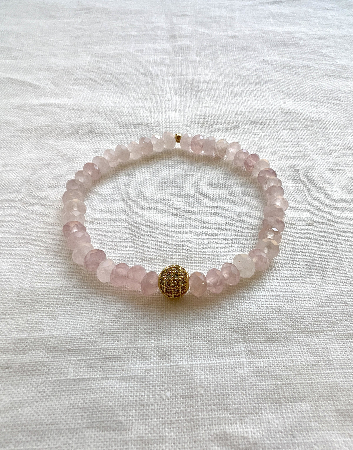 Faceted rose quartz rondelle stretch bracelet with a single gold plated pave CZ bead, laid flat on white linen