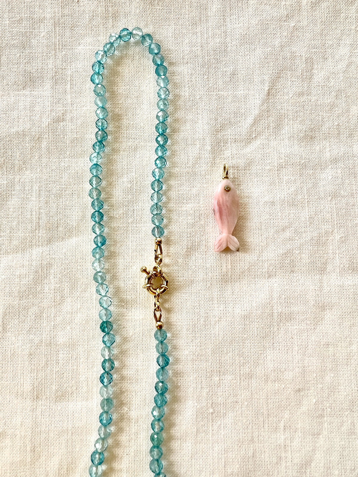 A beaded necklace made of faceted light blue topaz beads with a gold-filled sailor clasp with a removable pink opal fish charm