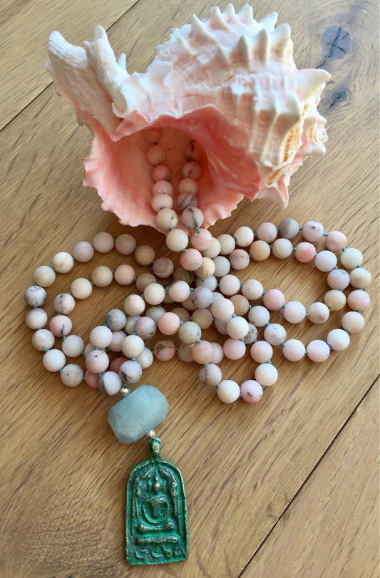 Pink Peruvian opal mala on a wood floor with a conch shell, a carved green Buddha pendant below an aquamarine nugget