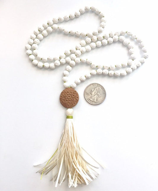 White lava stone mala with a terracotta diffuser disc and a cream raffia tassel, a US quarter for scale