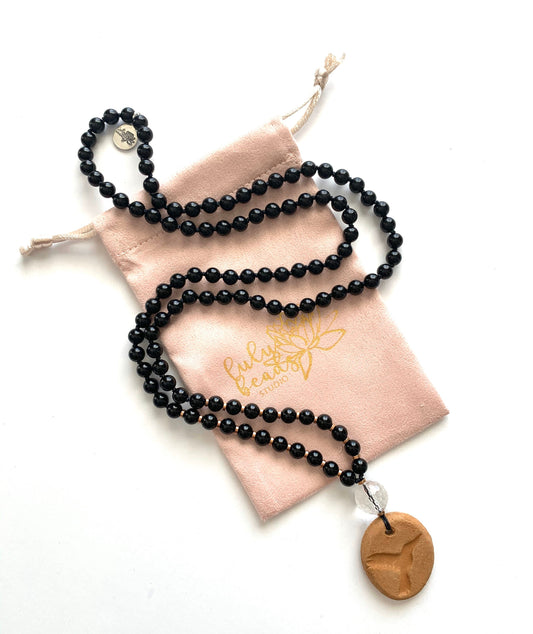 Black onyx mala curving across a blush pouch with a terracotta hummingbird diffuser pendant
