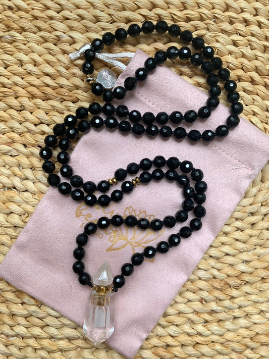 Faceted black onyx mala on a blush pouch and woven mat with a clear quartz perfume bottle pendant