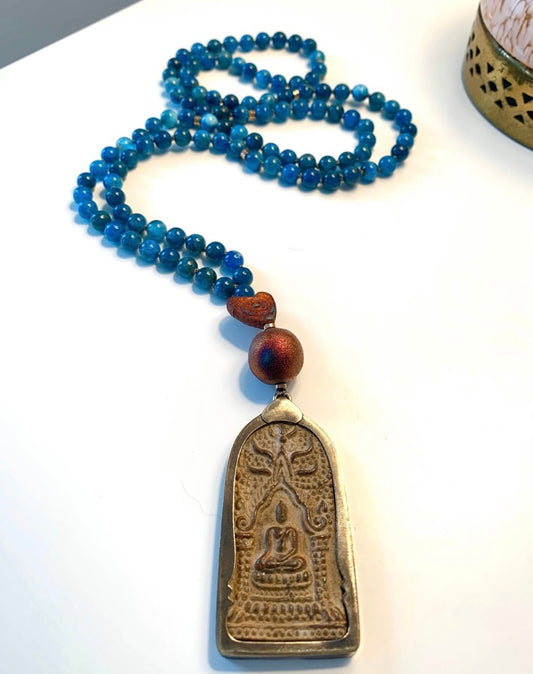 Blue apatite mala laid in a long S curve on white, ending in a raku pottery Buddha pendant